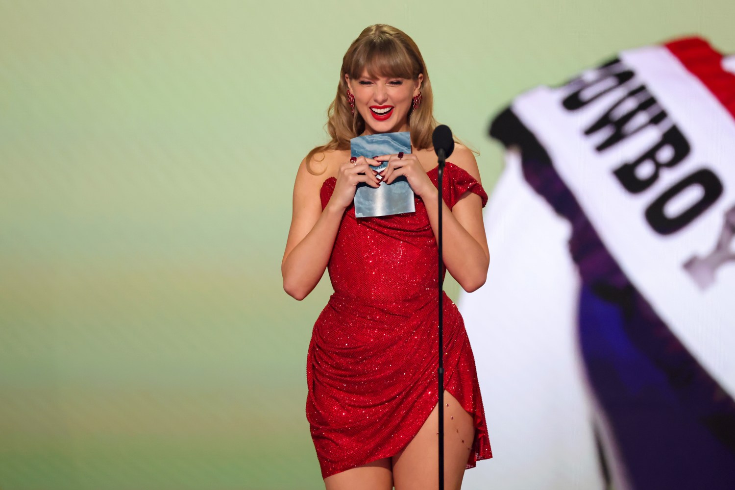 Taylor Swift stands on stage during the Grammy Awards in a red dress clutching an envelope.